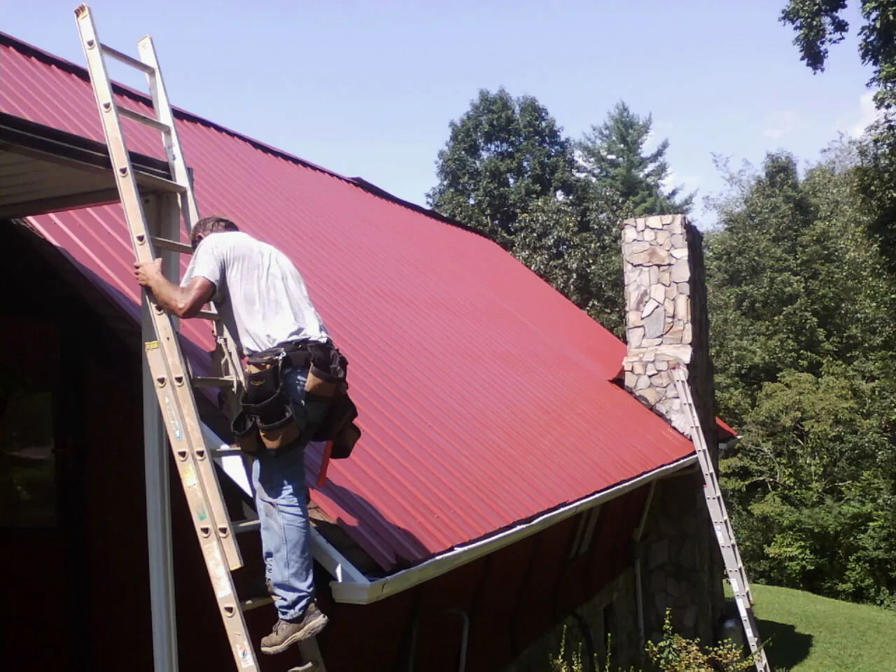 Roofer on a ladder beside a red metal roof and stone chimney — Soffit & Fascia Repair by Roofing Brothers in Crocheron